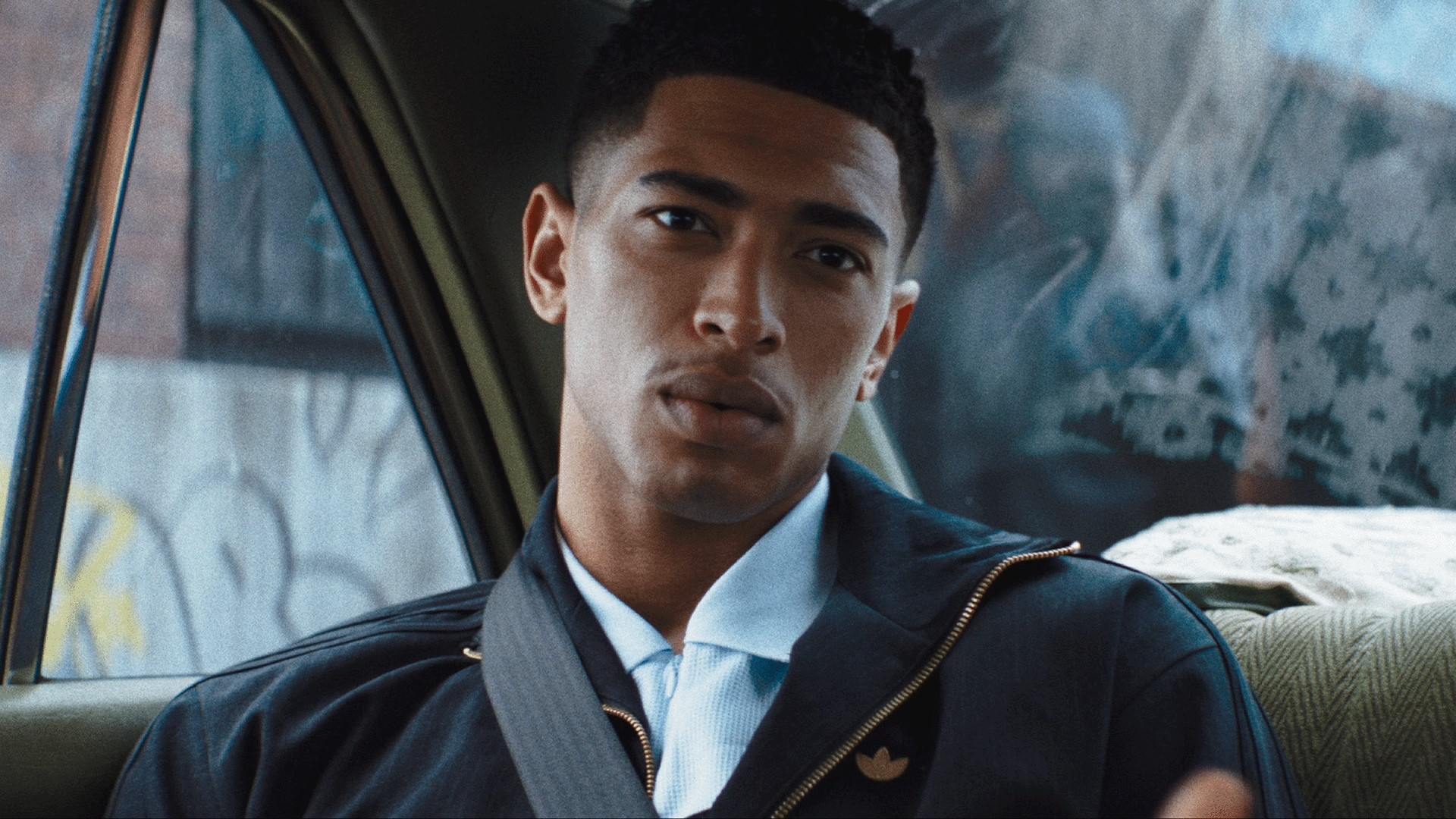 Jude Bellingham sits in the back seat of a car, wearing an adidas jacket and looking toward the camera.