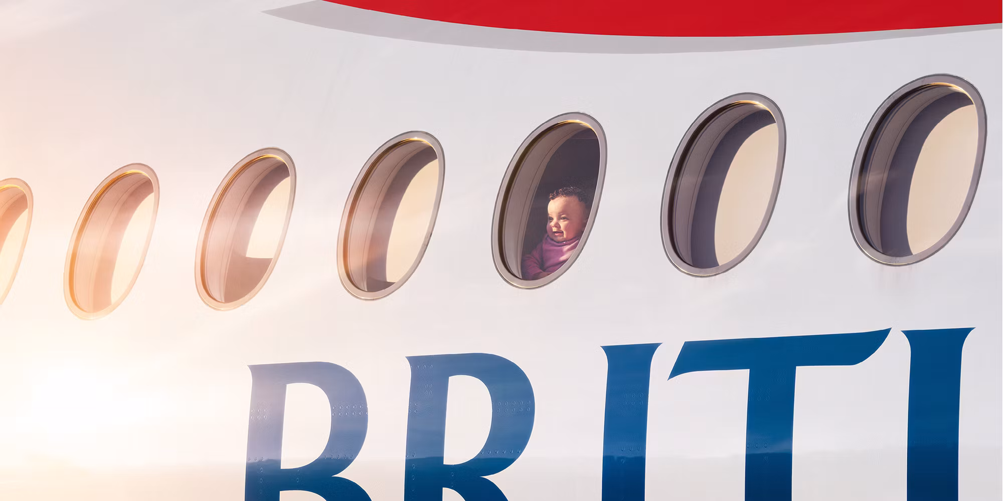 A young child gazes from one window of a warmly lit British Airways aircraft, with only cropped fragments of the red Speedmarque and blue wordmark visible.