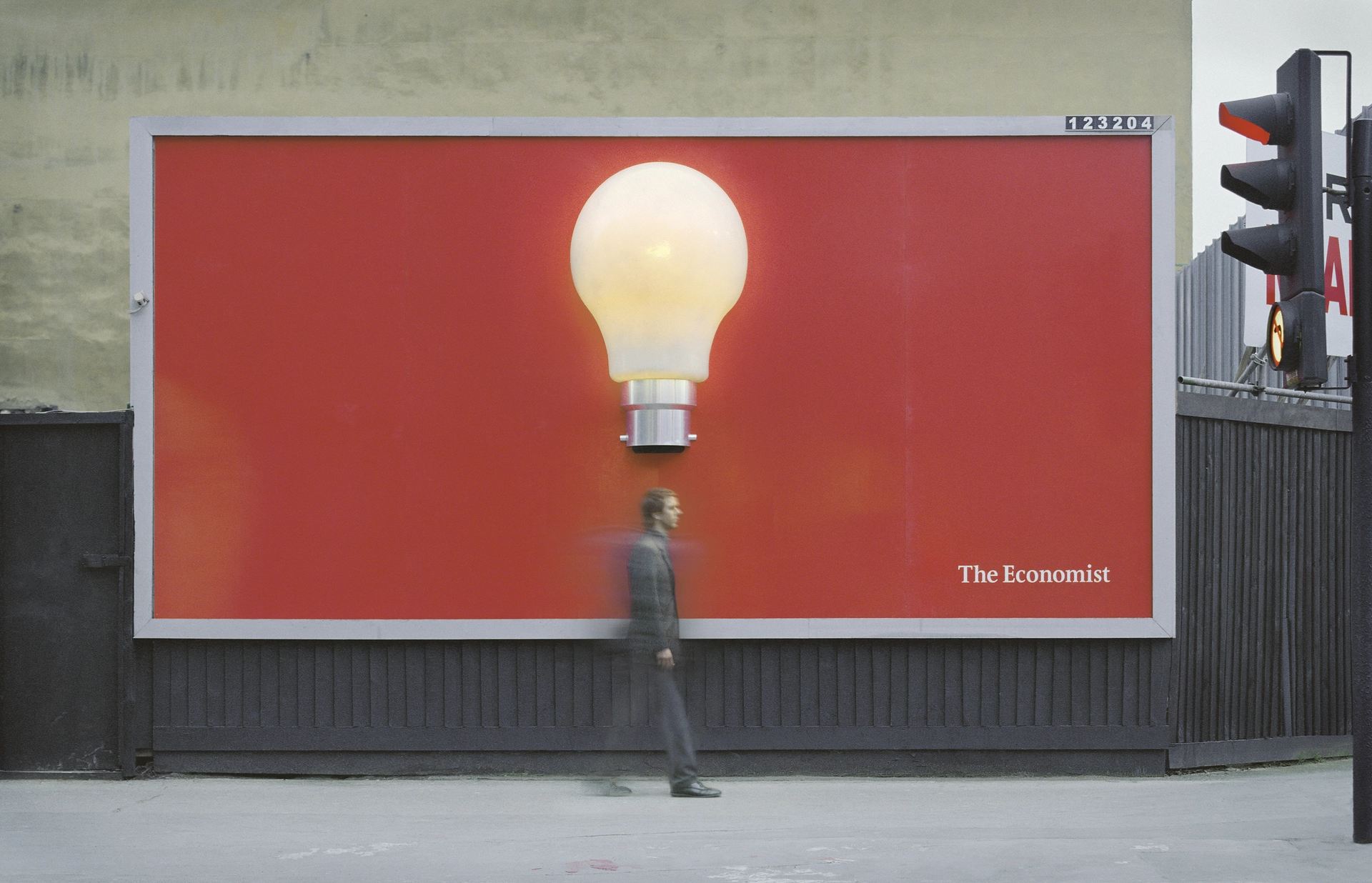A passerby walks beneath an illuminated three-dimensional light bulb mounted on a red Economist billboard.