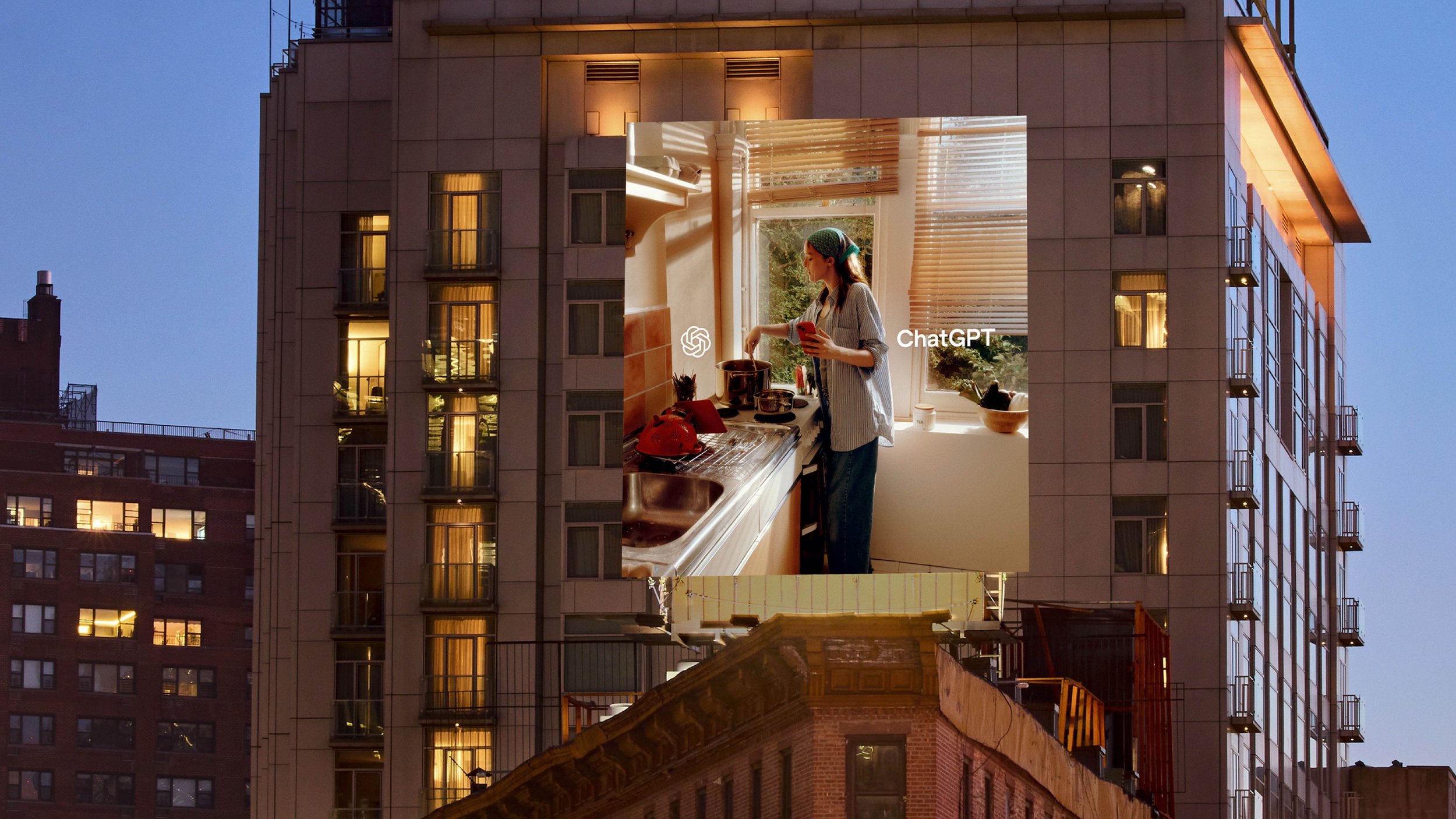 A tall ChatGPT billboard showing a person cooking in a sunlit kitchen is installed on a city building at dusk.