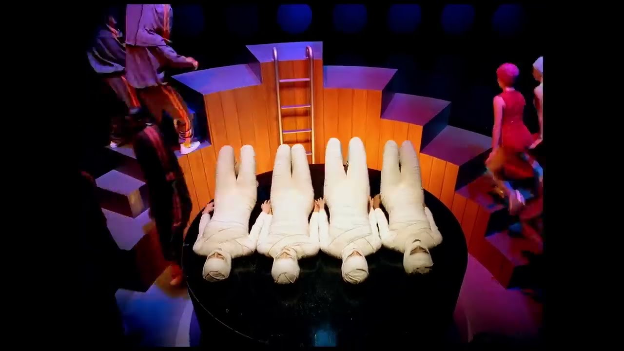 Costumed dancers arranged around a circular stage, with four white-clad figures lying in its centre, in Around the World.