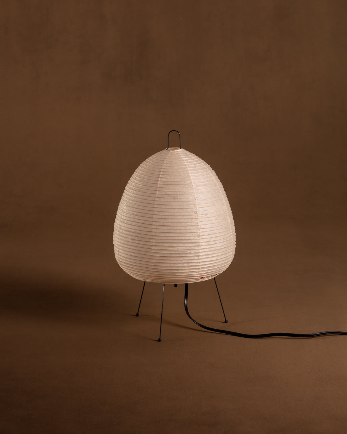 An unlit Akari 1A lamp with a rounded ribbed paper shade on three thin metal legs against a warm brown background.