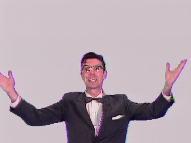 David Byrne in a dark suit and bow tie, raising both hands against a pale background in Once in a Lifetime.